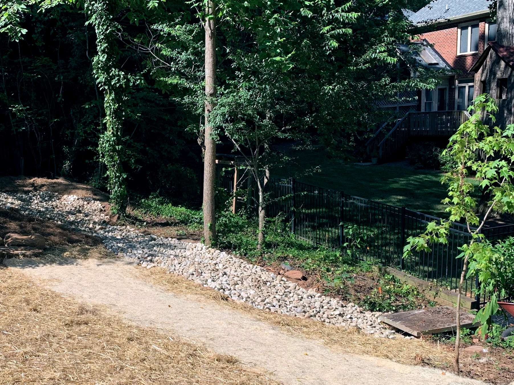 Residential backyard with drainage solutions including a river stone trench and covered drain in Winston-Salem, with a green lawn and wooded area in the background.