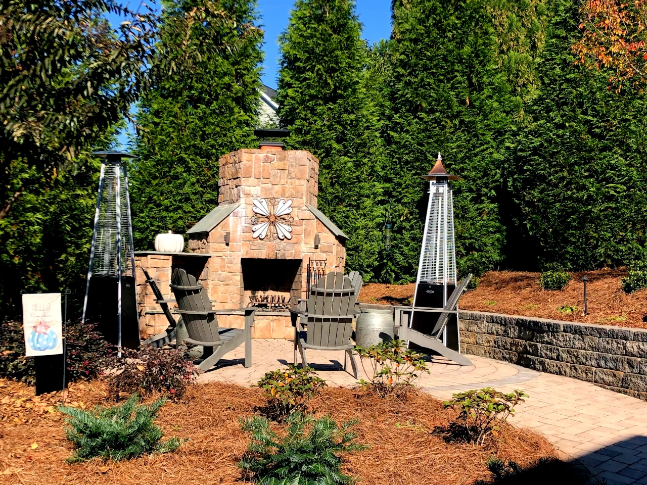 Beautifully designed outdoor hardscape with a stone fireplace, patio heaters, Adirondack chairs, and surrounded by evergreen trees at a Winston-Salem NC home.